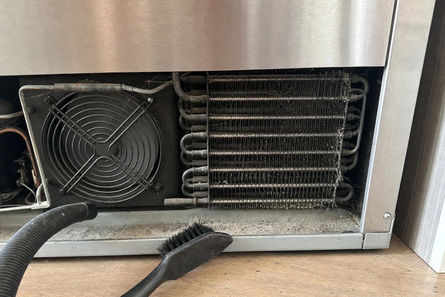 Dust-matted condenser coil and condenser fan behind the lower grille of a stainless built-in, vacuum hose alongside
