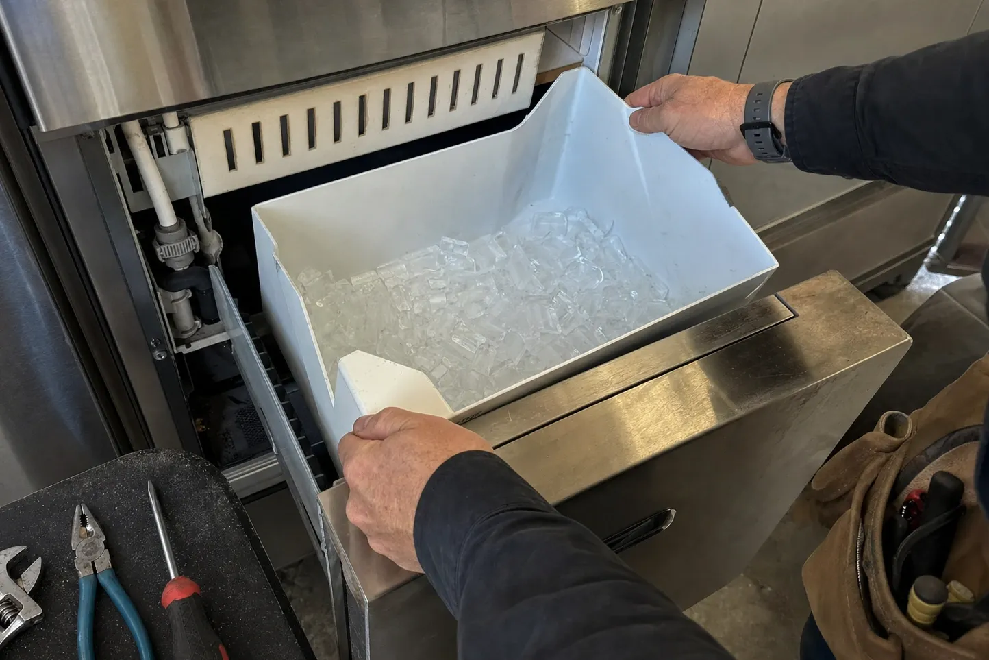 Hands lifting a bin of clear cubes out of an undercounter ice maker, pliers and drivers on a floor mat