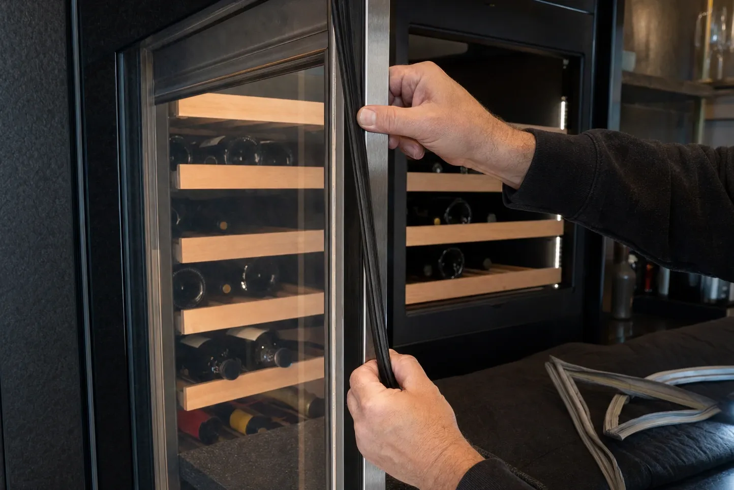 Hands fitting a door gasket along the glass door of a wine cabinet, bottles resting on wooden racks behind